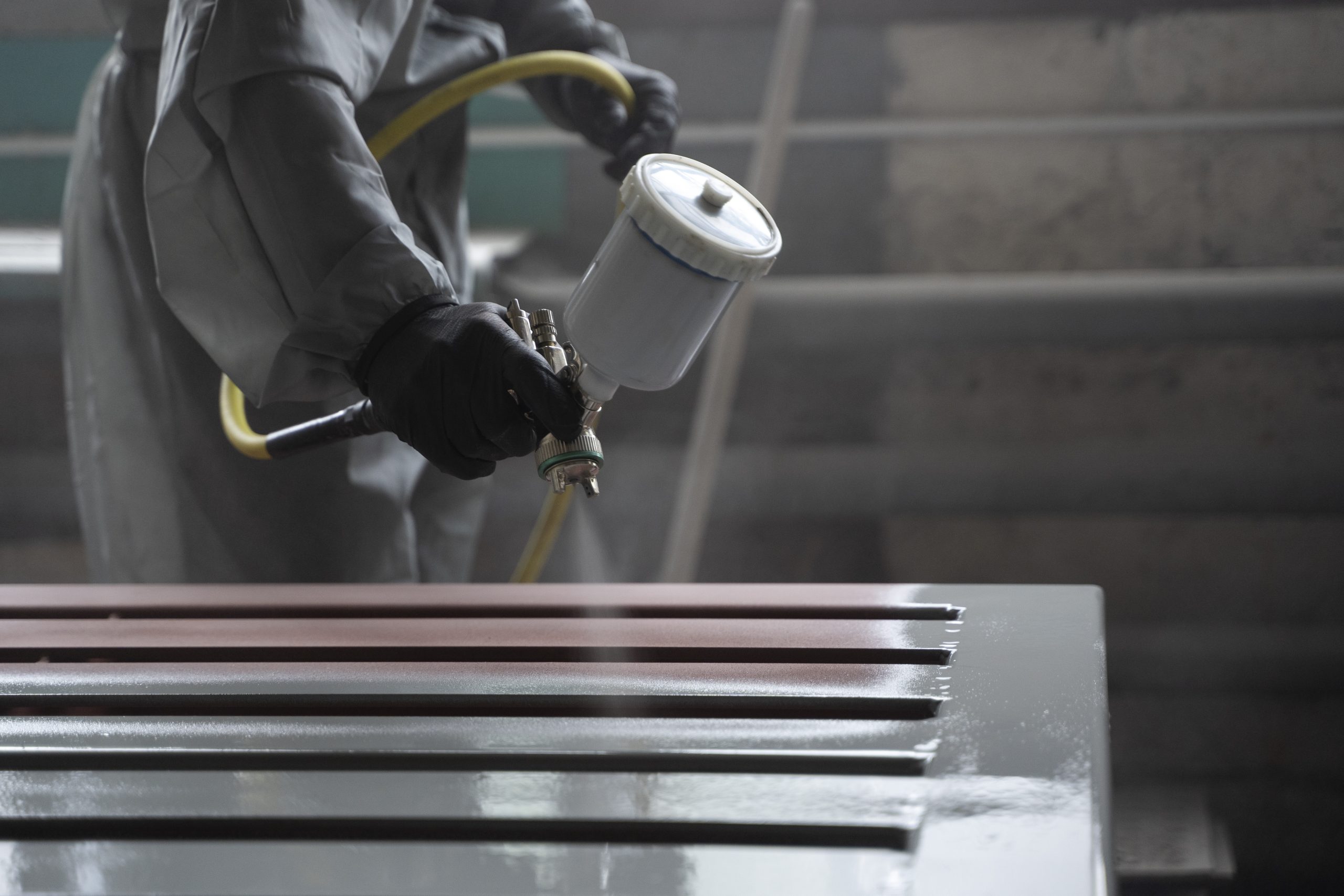 Close-up de um profissional industrial, usando luvas pretas, aplicando tinta cinza com uma pistola de pulverização sobre uma superfície metálica. Parte da superfície já está pintada, enquanto outra mostra a cor original ou uma camada de primer marrom avermelhado. Fumaça ou névoa de tinta é visível ao redor do bico.